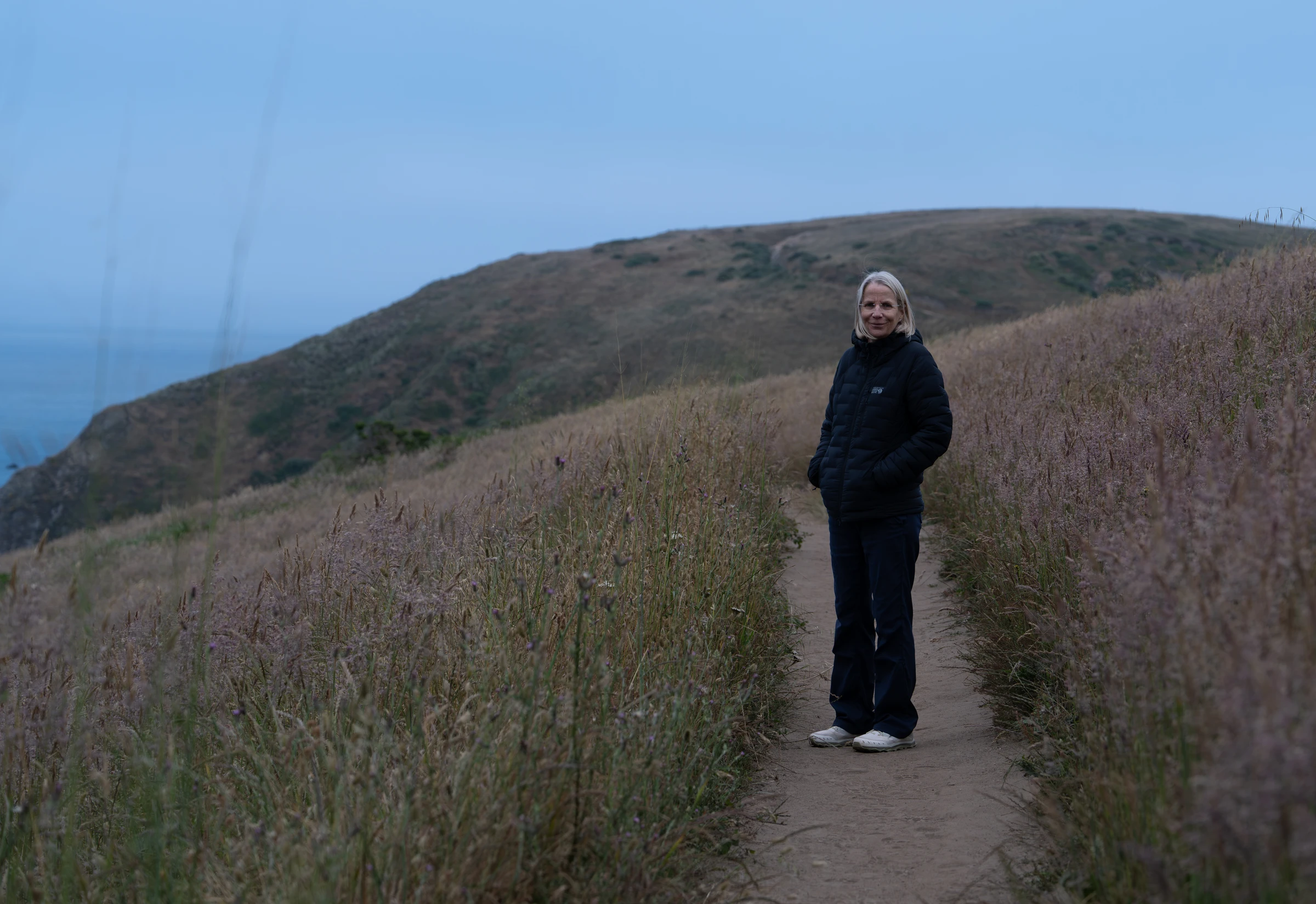 Britta Butler standing on a grassy coastal hillside beneath a blue sky.
